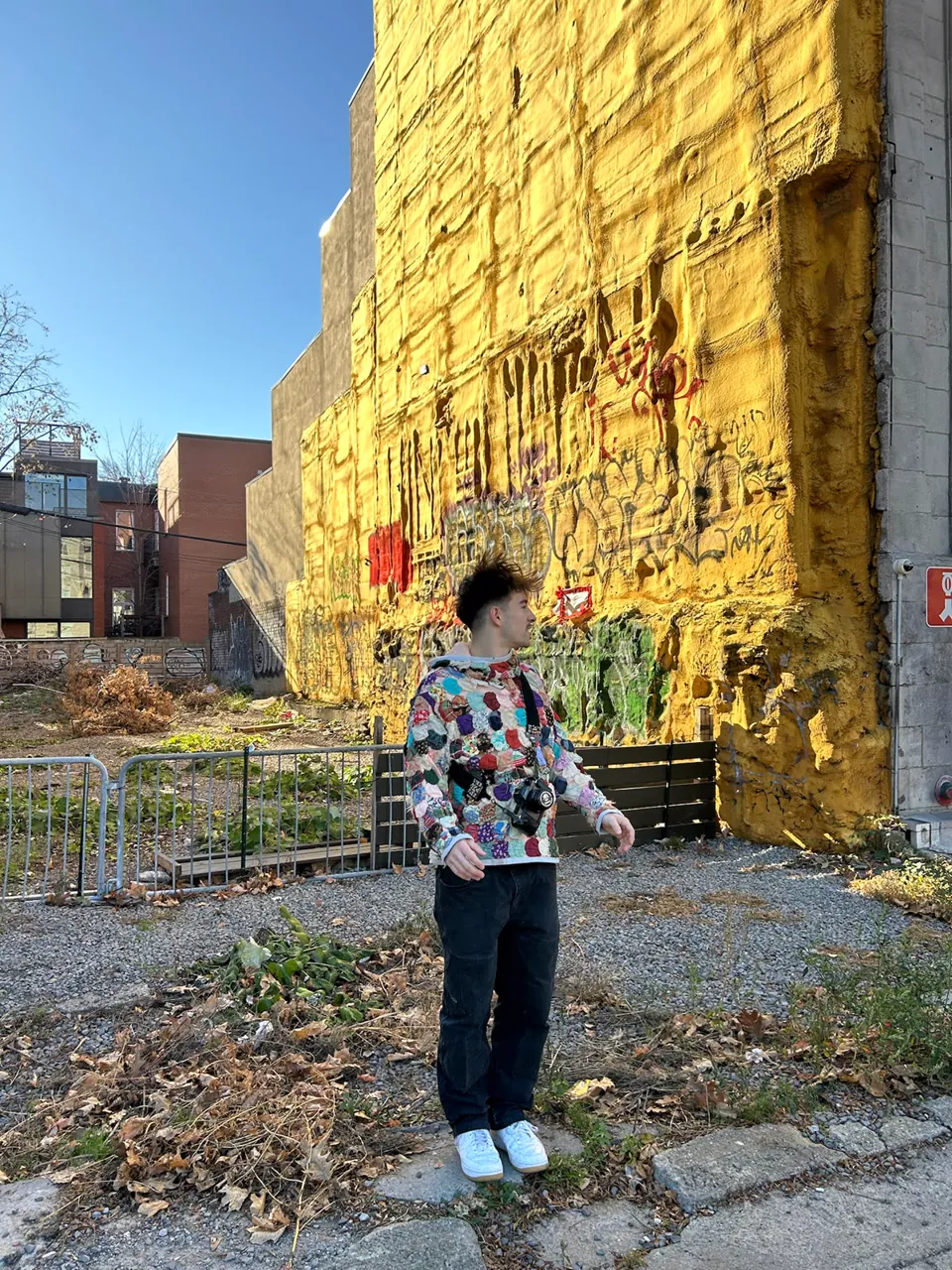 Hoodie covered edge to edge in multicolored quilt yo-yo rosettes, worn in front of a yellow graffiti wall