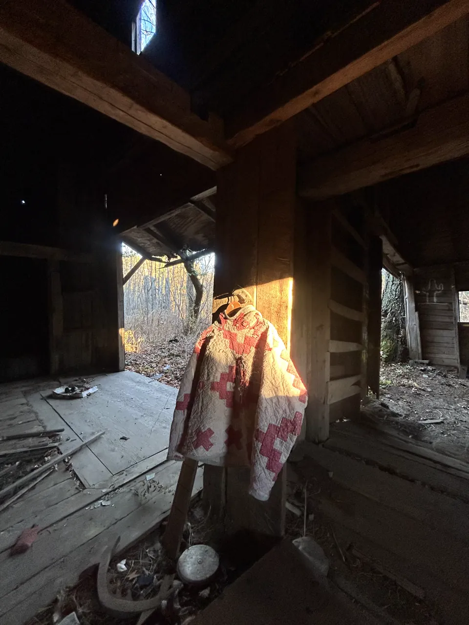 Red and white cross quilt pullover hanging in a dark barn, lit by a shaft of low sun