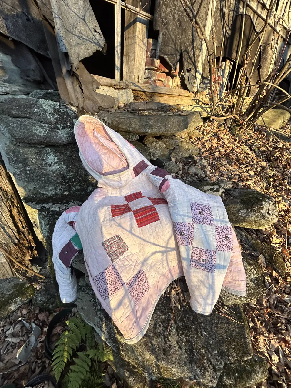 Pale pink nine-patch quilt hoodie draped over mossy stones by a collapsing barn