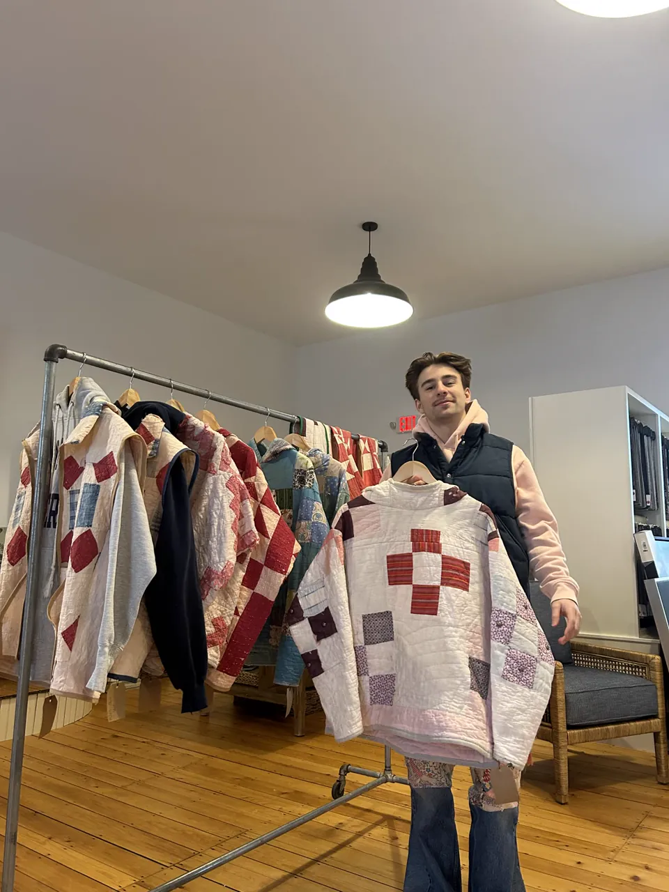 Sam holding a white and red quilt pullover on a hanger, in front of a rack of quilt garments